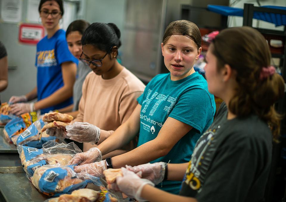 Students wearing hairnets and gloves work in a line at a kitchen counter.