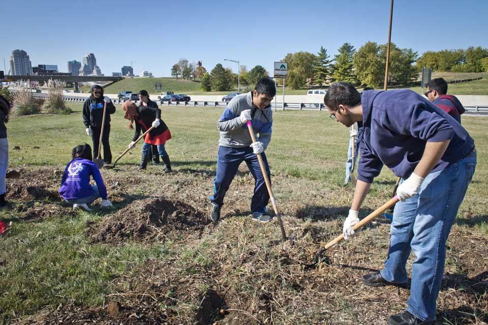 Students use shovels and other equipment in a grassy area next to a highway.