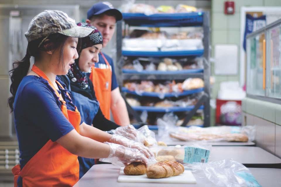 Three students are shown in profile, wearing aprons, working with bread on a kitchen counter.