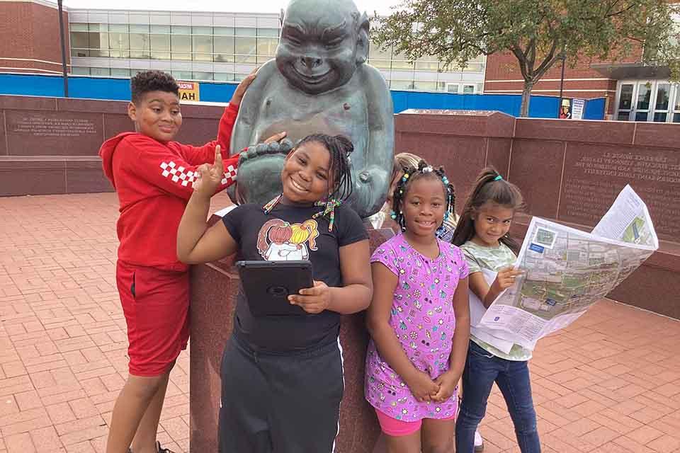 iScore students gathered around the Billiken statue, one holding a map, during a geocaching exercise