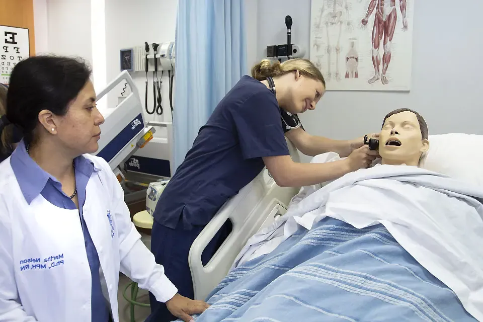 一名穿着医疗服的学生在医院病床上检查病人模型的耳朵，而一名教师在旁边观察.
