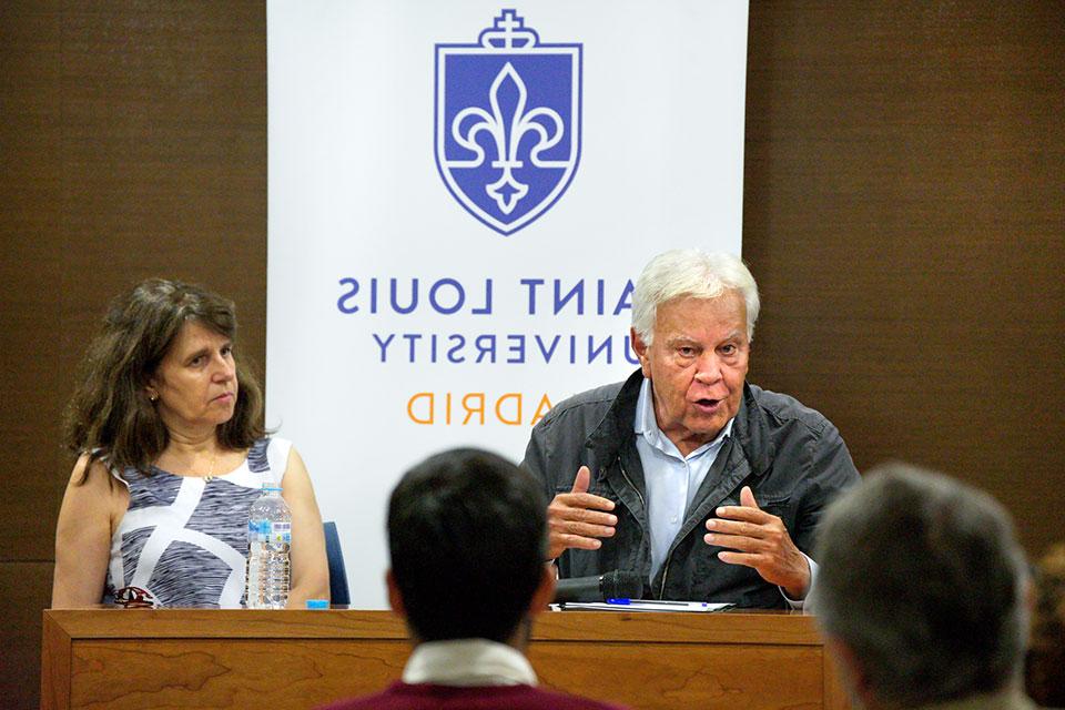 Dr. Laura Tedesco sits next to former Spanish Prime Minister Felipe González during a discussion. They sit at a wooden table in a lecture hall.