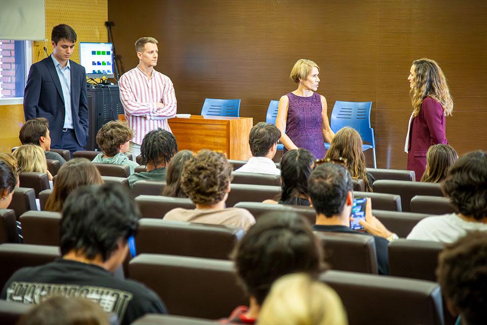 Four presenters share a discussion at the front of an auditorium with students, seen from behind, in the audience.