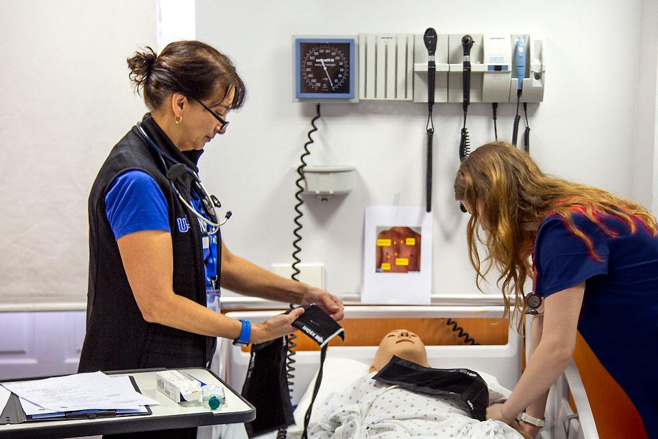 一名教师站在病床的一侧，病床上放着一个拿着血压袖带的病人模型，而另一侧则有两名学生在观看.