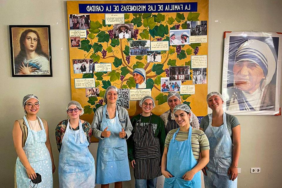 A group of seven students wearing aprons and hairnets pose in front of a large painting that reads La Familia De La Misionera De Caridad.