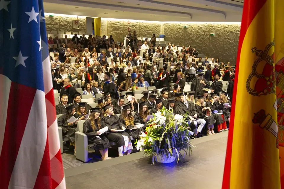 Spanish and U.S. flags frame an image of graduates and sitting in an auditorium wearing caps and gowns and their families.