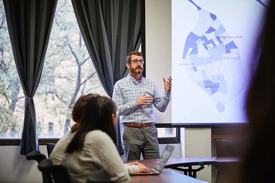 CCJ program director Joseph Schaeffer stands in front of a classroom giving a presentation with a St. Louis map on the screen.