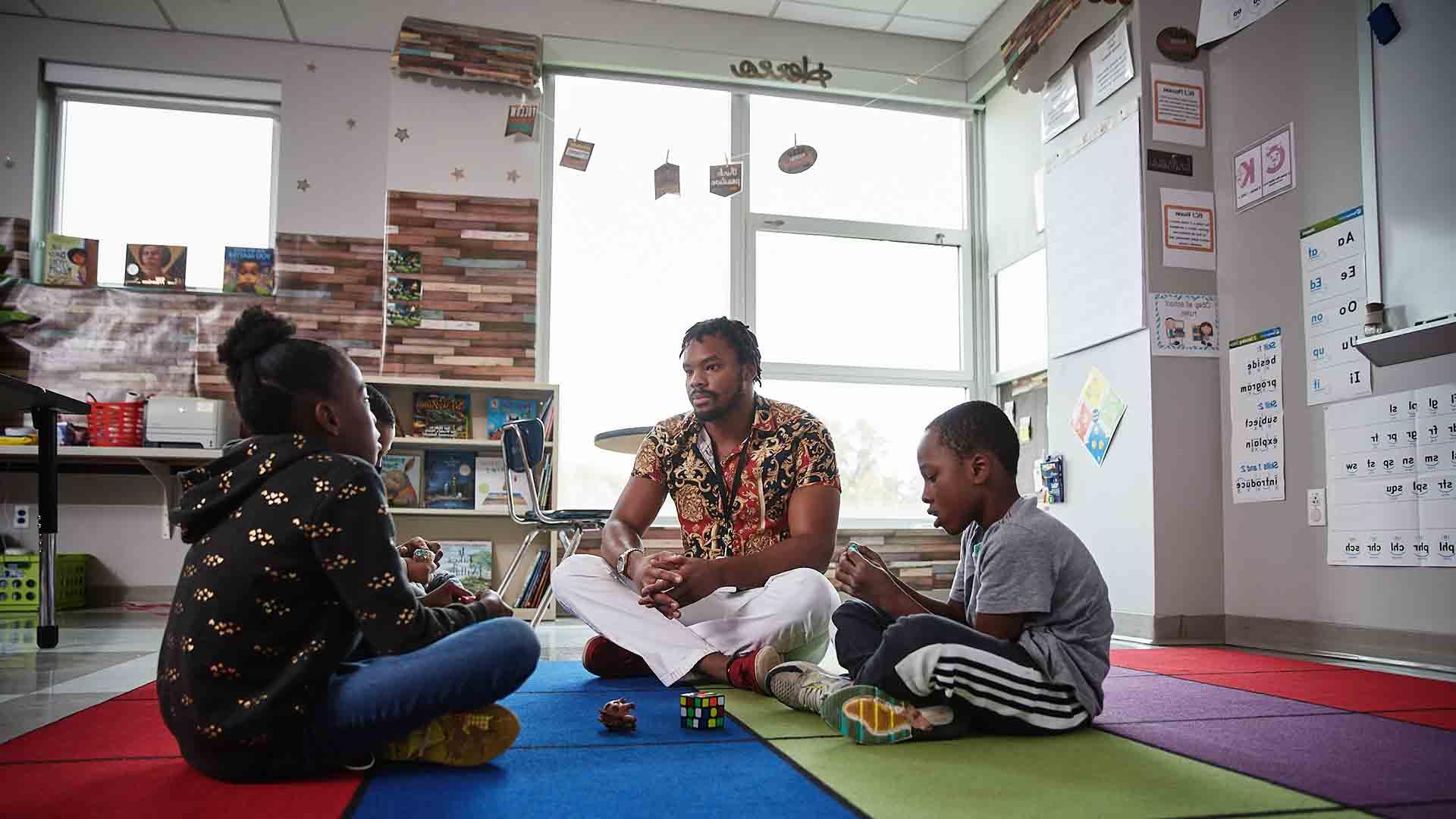 A School of Social Work student sits on the floor of a colorful classroom, talking to two young children.